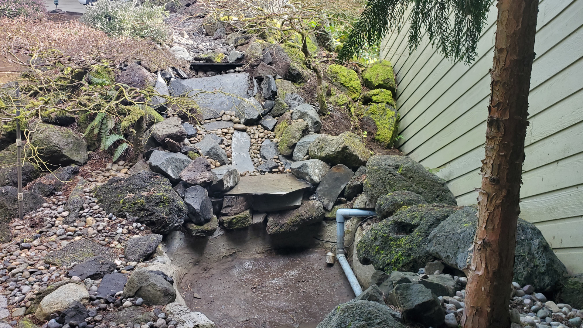 A steep, short waterfall now covered in various rocks. Large rocks line the edges with flagstone slates mixed into the center and river rock covering all of the liner.