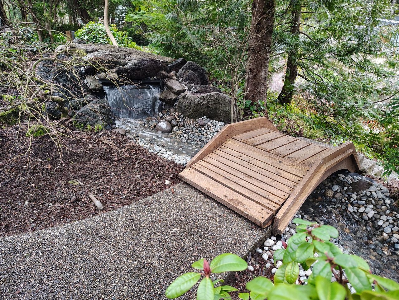 A stream covered with river rock. A water fall spillway is about 2 feet off the ground, cascading water onto the rocks. There is a concrete pathway with a small wooden bridge that crosses the stream.