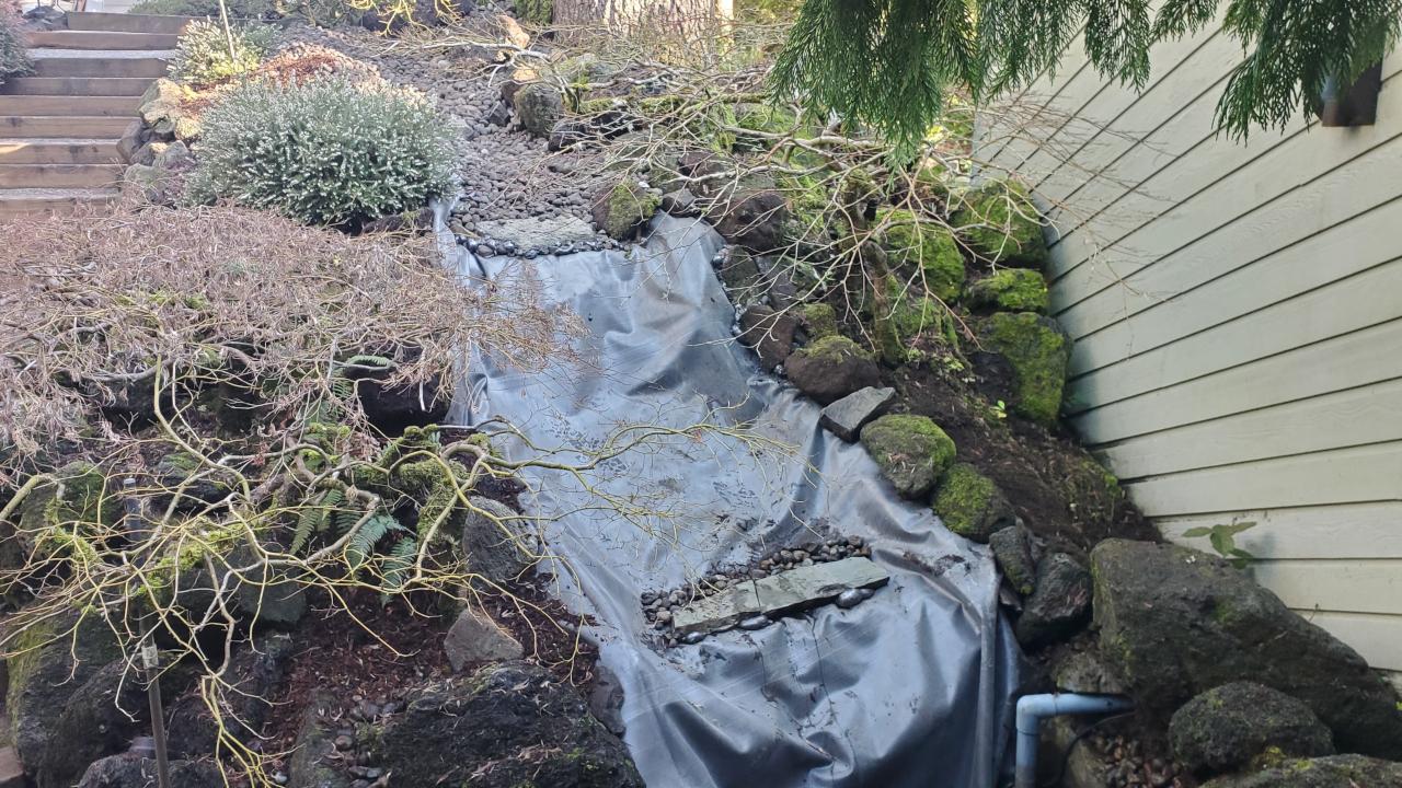 A steep, short water fall with the bare liner showing. The edges are lined with large rocks and plants